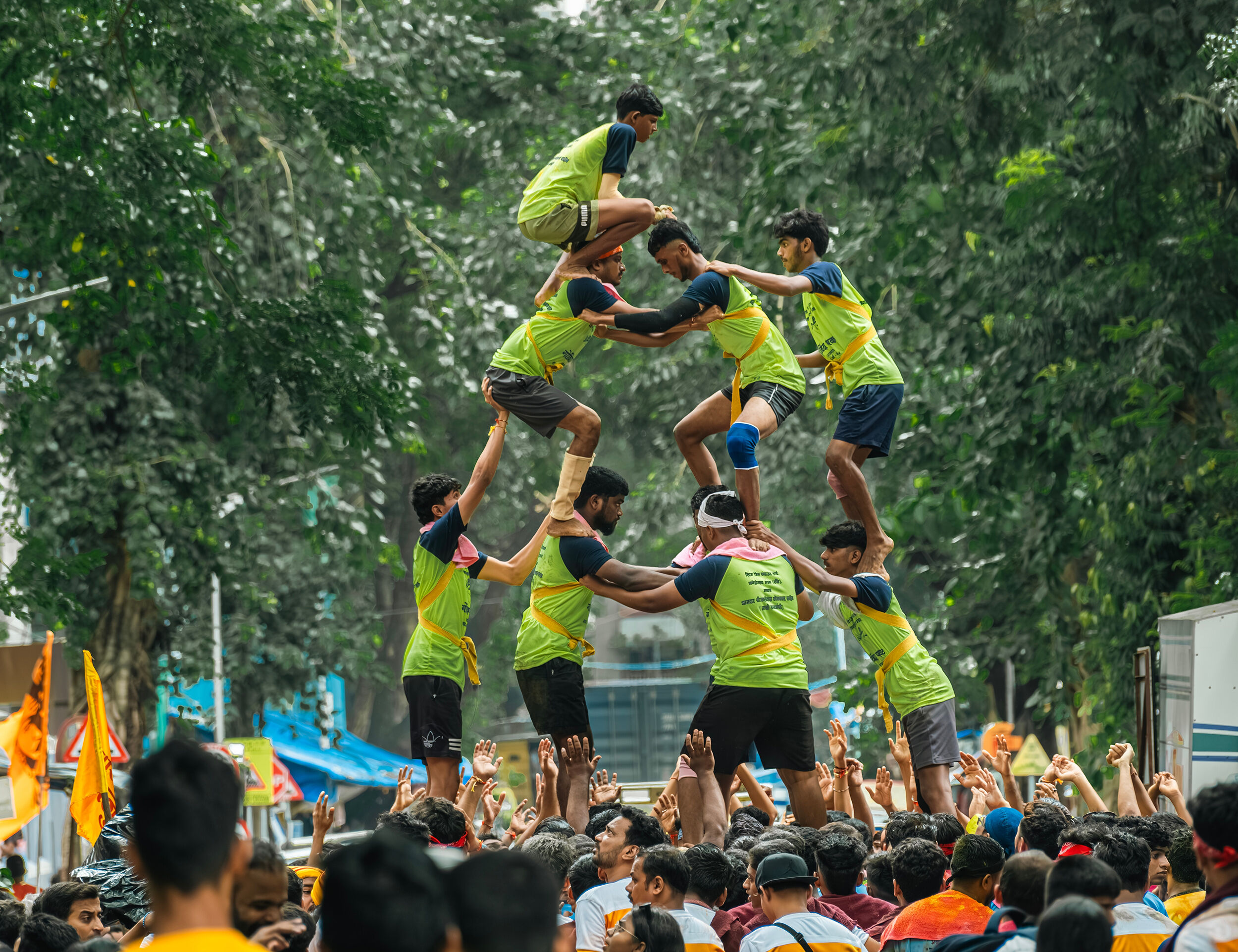 Group of young men forming a human pyramid during the Dahi Handi celebration on Janmashtami, with a cheering crowd and greenery in the background.